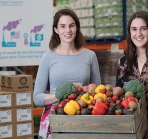 Two women standing behind a wooden box filled with fresh vegetables