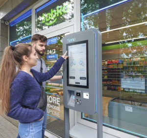 Two shoppers stand outside a Zabka store and enter their bank card into the entry portal to gain access to the store.
