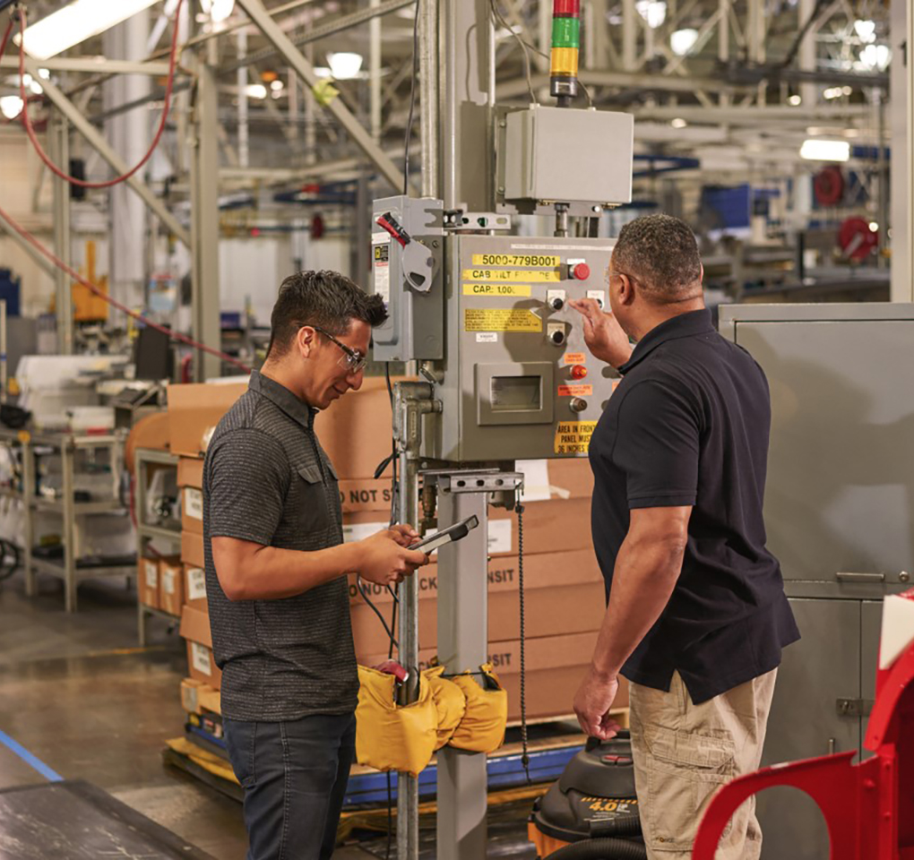 Two men on a factory floor, one interacting with a tablet device and the other punching buttons on an electrical panel.