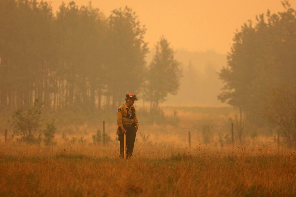 A firefighter stands in a field amidst dark, heavy smoke in the air