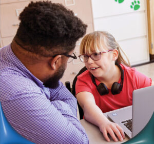 Girl in a classroom looks up at her teacher while working on a laptop computer