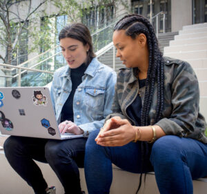 Two women sitting on stairs outside a building and talking about what's on a laptop computer one of them is holding