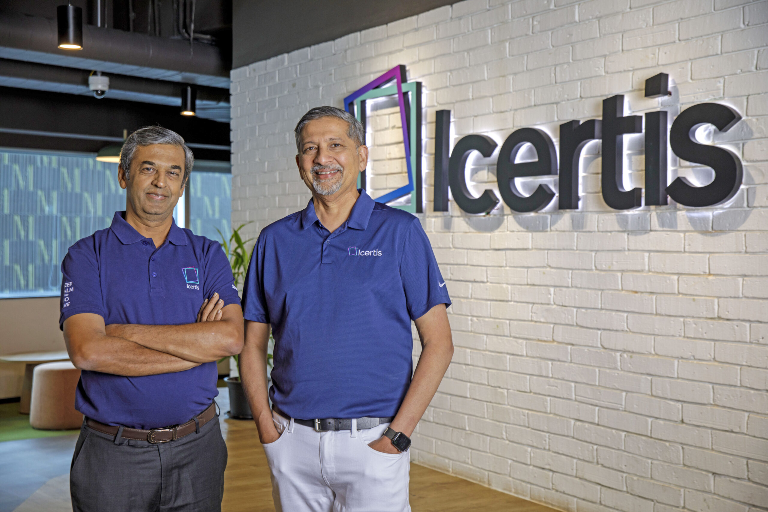 Two men stand in front of a white brick wall with company name Icertis behind them.