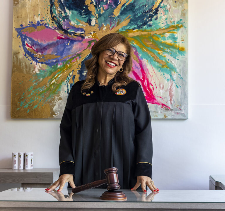 A female judge in robes stands at a desk with a gavel in front of her, behind her a colorful painting