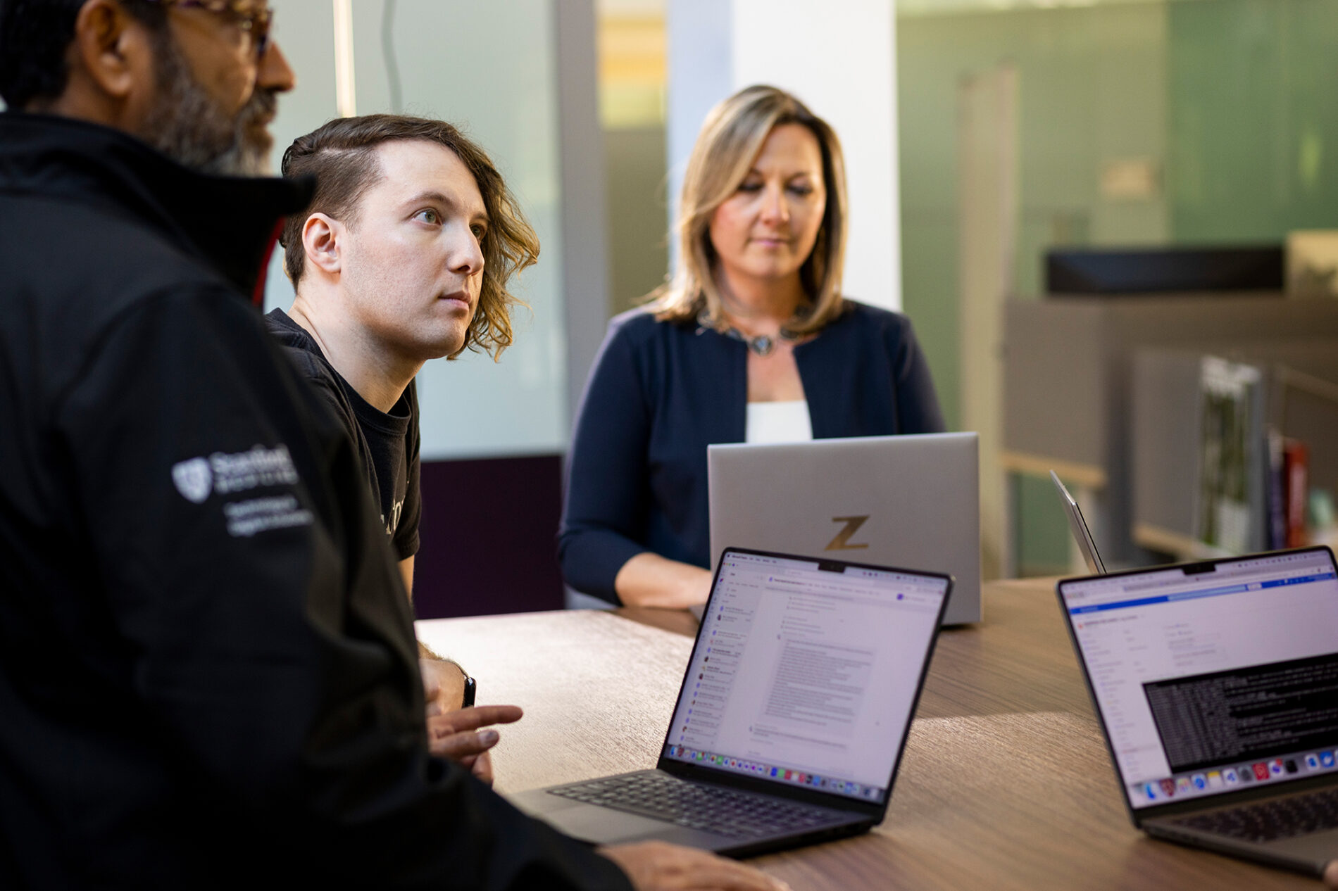 A man with an asymmetrical haircut, wearing a black T-shirt that says Stanford Medicine, stands with arms crossed in a glass office space