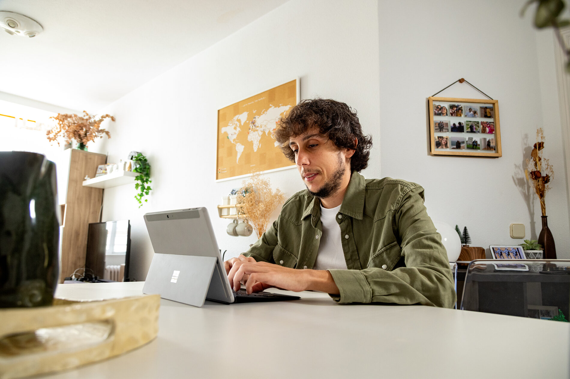 A man with dark, curly hair and a short beard, wearing a khaki shirt open over a white shirt, works on a laptop.