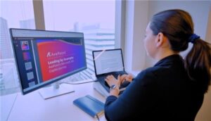 A woman sits at a desk in front of a window, typing on her laptop.