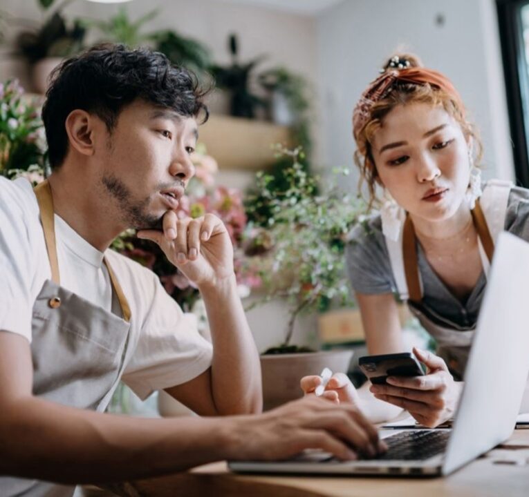 Two retail workers wearing aprons look at a laptop screen
