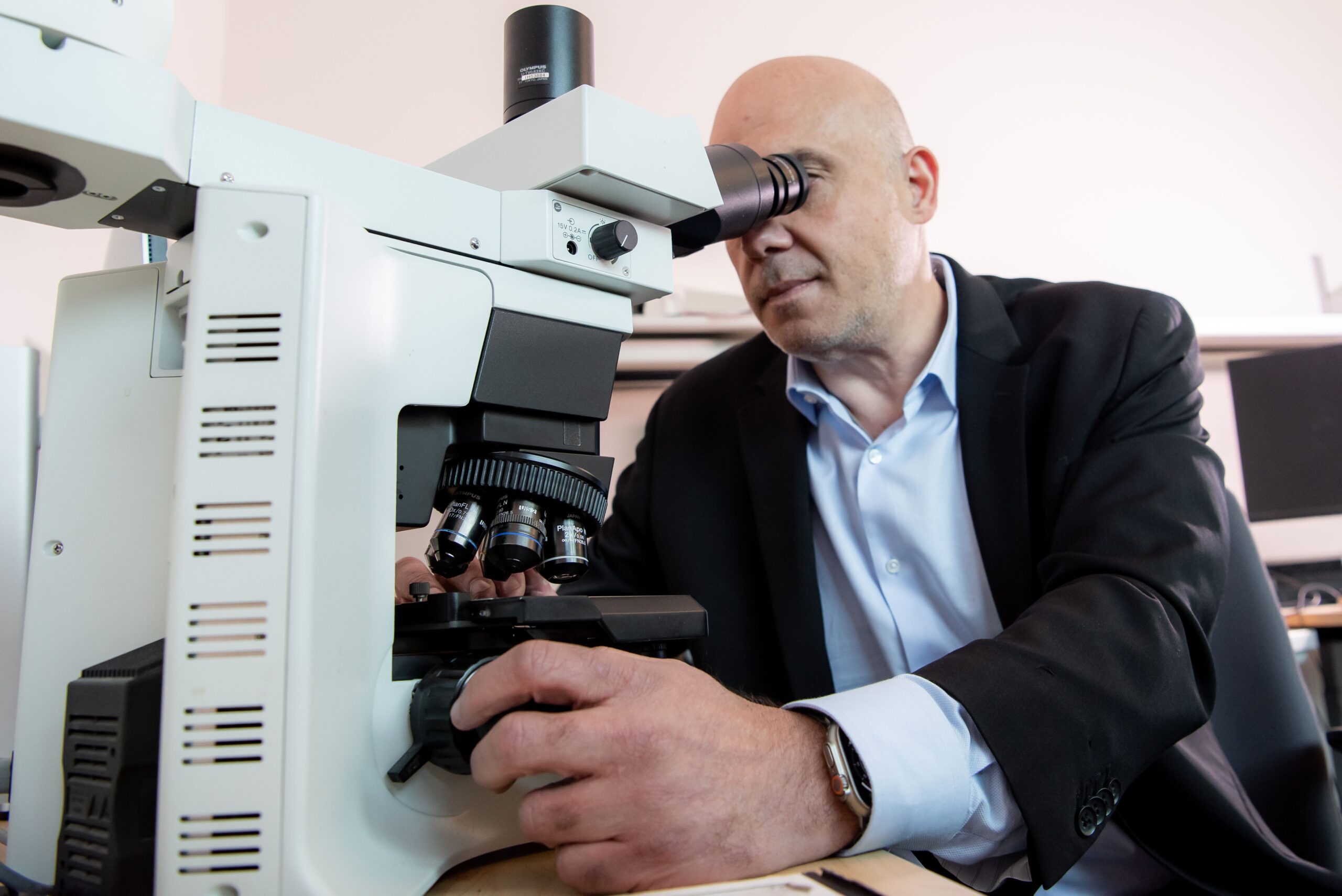 A person in a suit looks through a microscope, adjusting the focus knob while seated at a lab workstation.