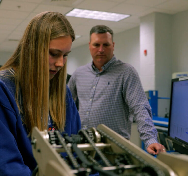 Teacher watching as a student performs on operation with a piece of machinery.