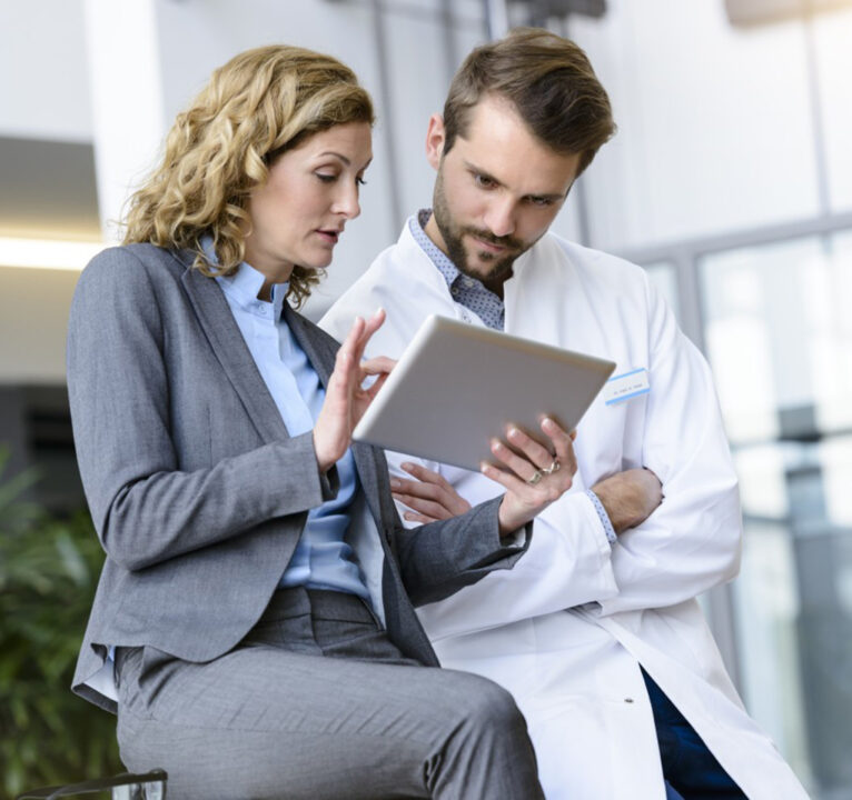 Two people, one wearing a white medical smock, discussing what's on the tablet device that one of them is holding.