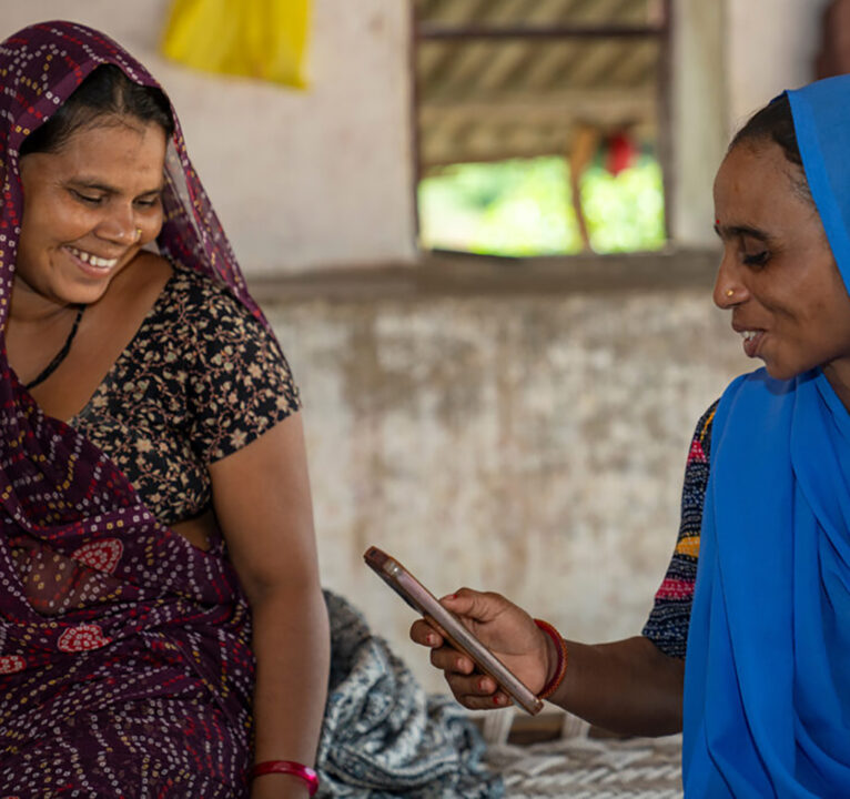 Two women in India smile as they discuss what's on the app of the phone one of them is holding.