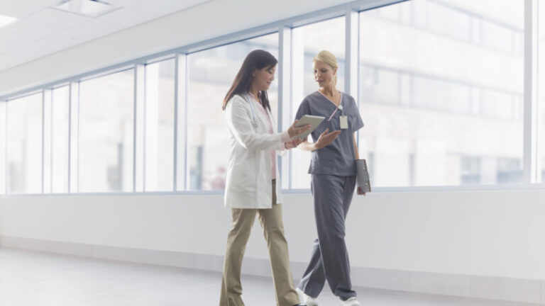Two healthcare professionals discussing what's on a tablet device as they walk down a hallway.