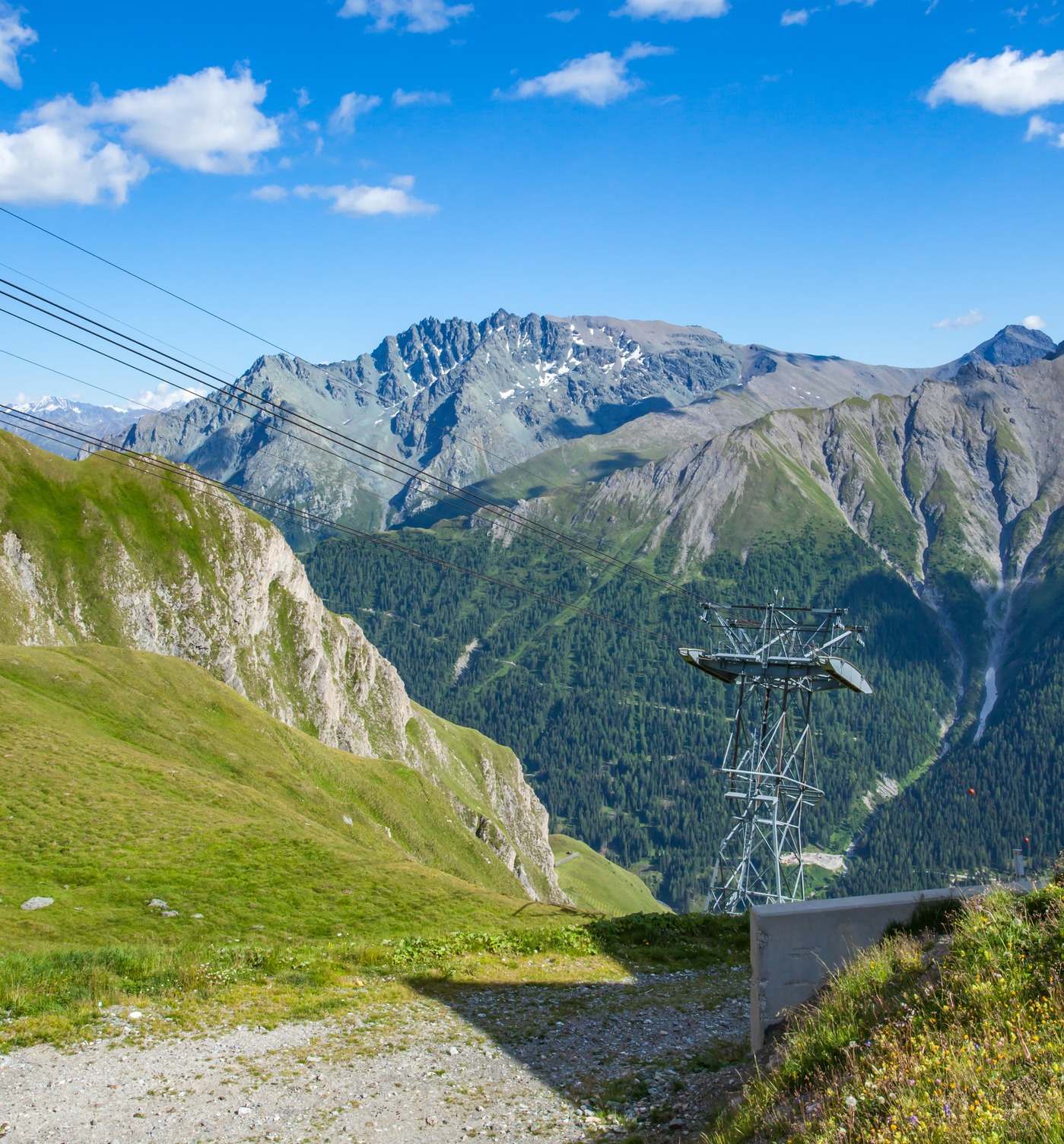 A cable car tower and overhead cables traverse a dramatic Alpine valley with snow-capped mountains, green meadows, and forested slopes under a blue sky with white clouds.