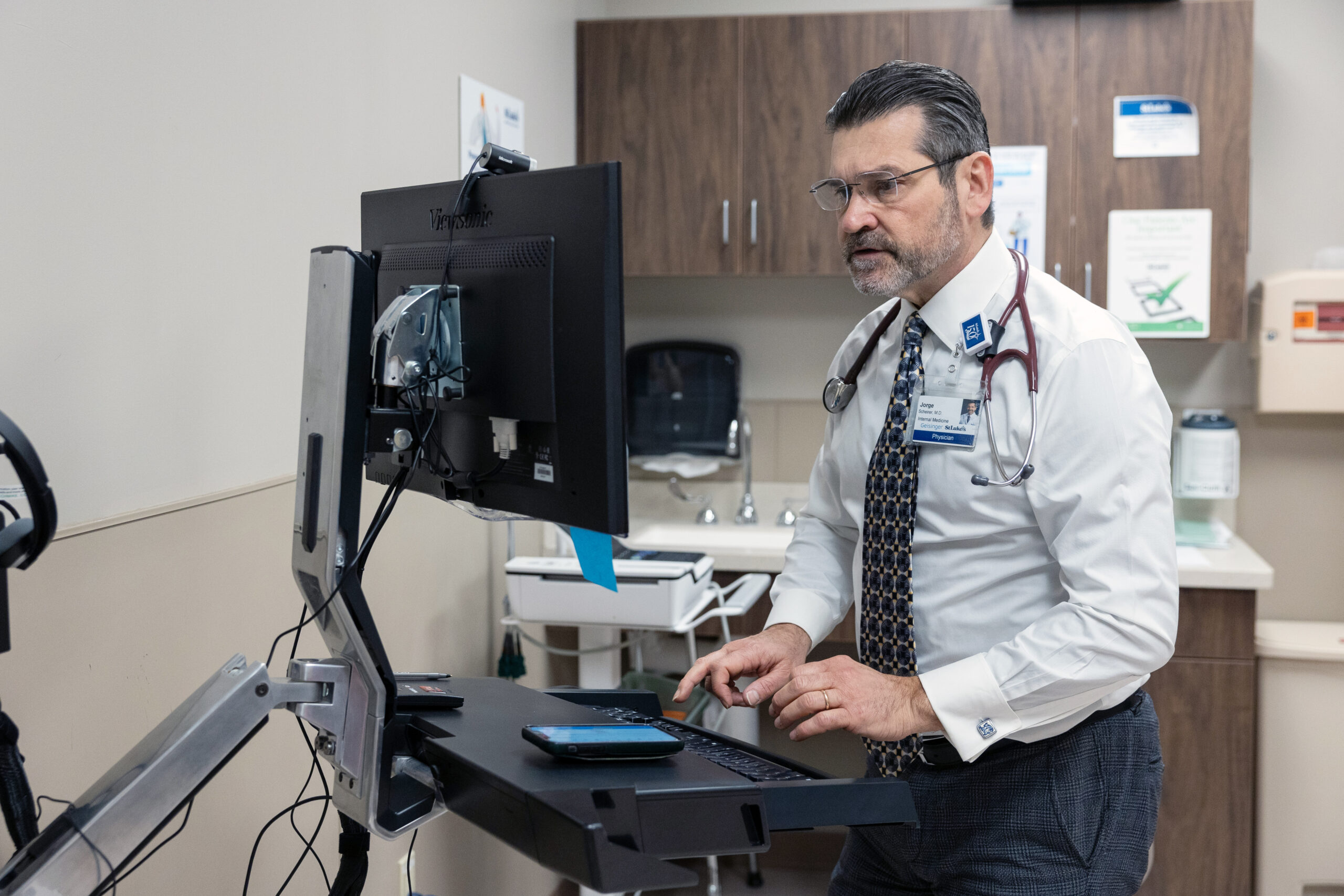 A doctor in a white coat with a stethoscope sits at a standing desk using a computer monitor in a medical office.