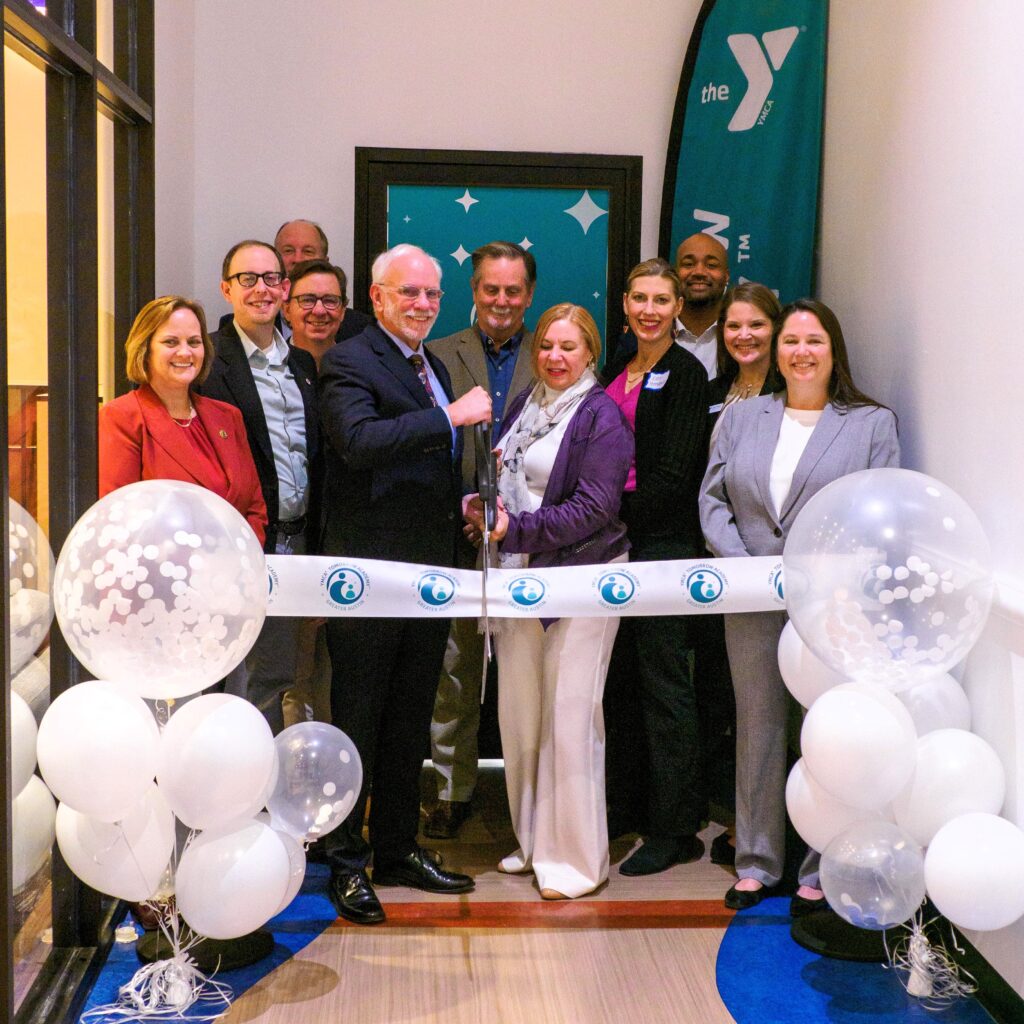 A woman surrounded by co-workers cuts a ribbon with a pair of oversized scissors to launch a new child care center.