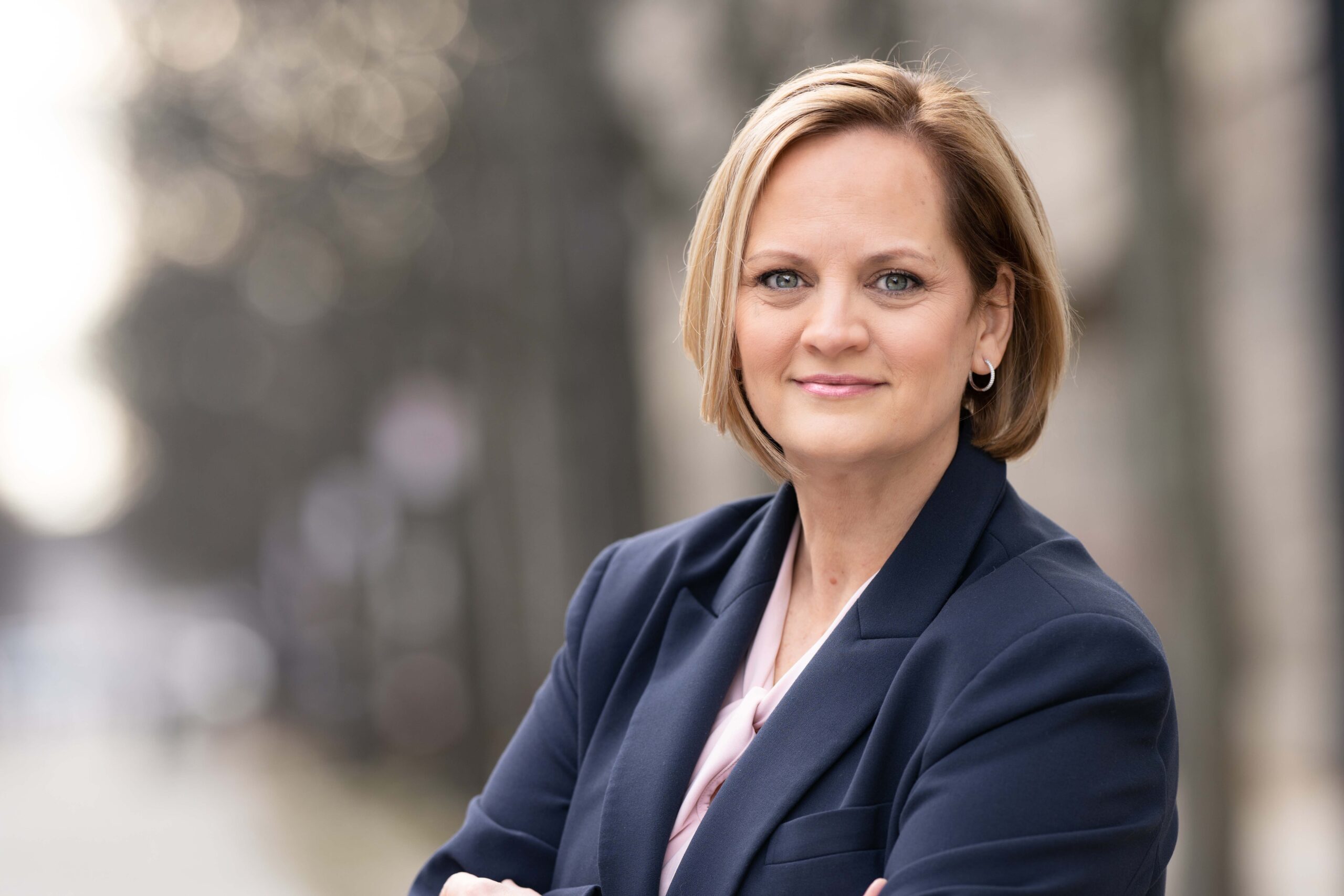 An individual with short, light brown hair wearing a navy blue blazer standing outdoors with arms crossed
