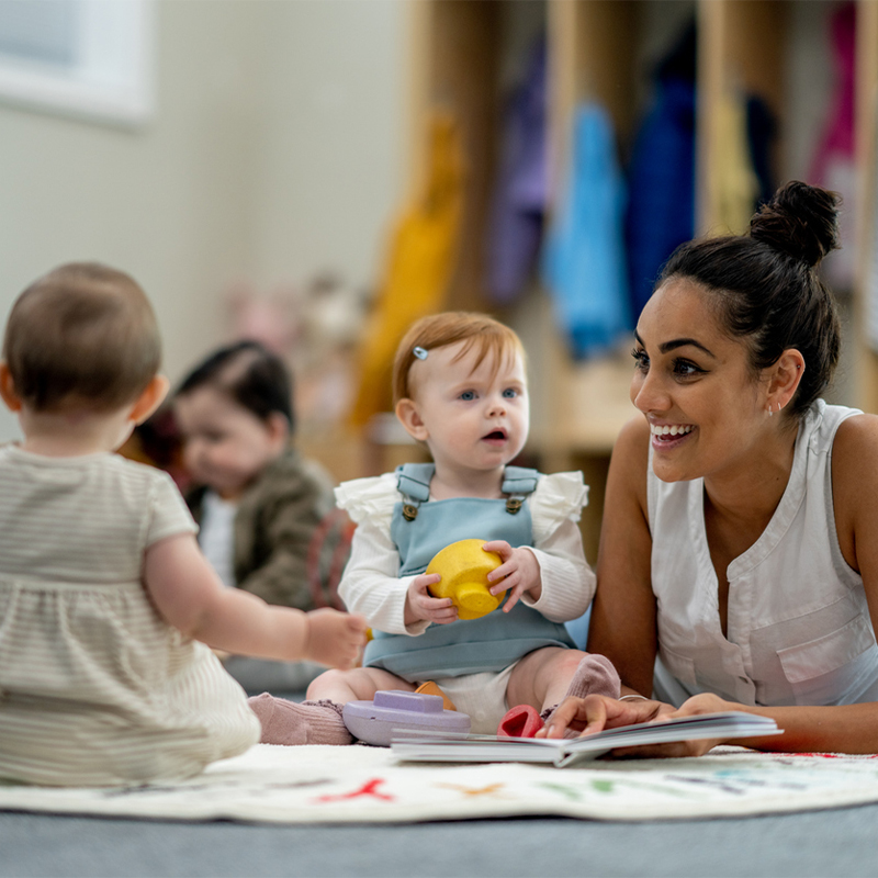 A group of young children and an adult sitting on a soft surface indoors, engaged in play and exploration.
