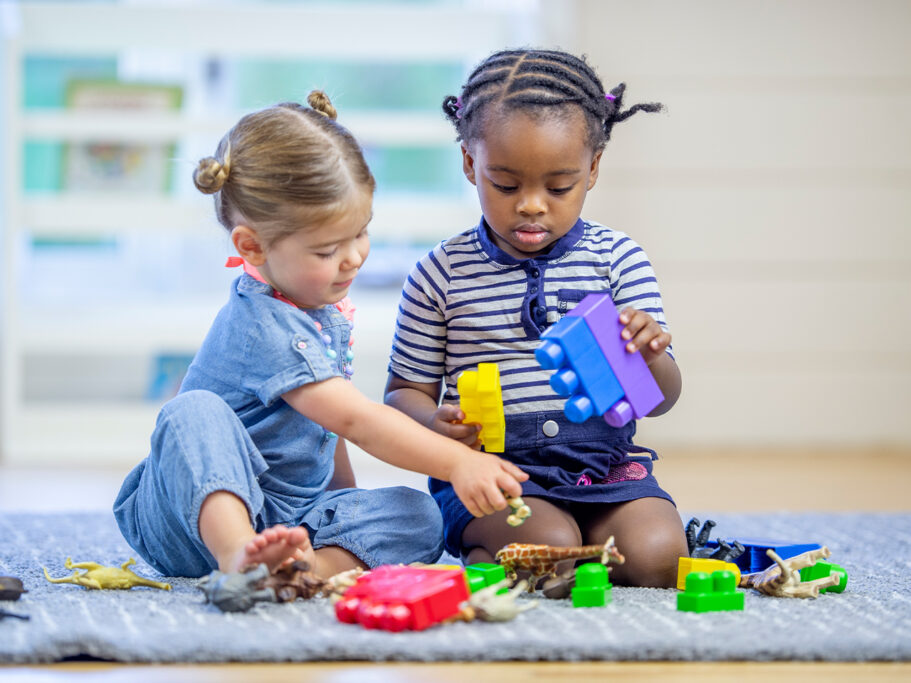 Two children sitting on a gray carpeted floor, playing with colorful toys.