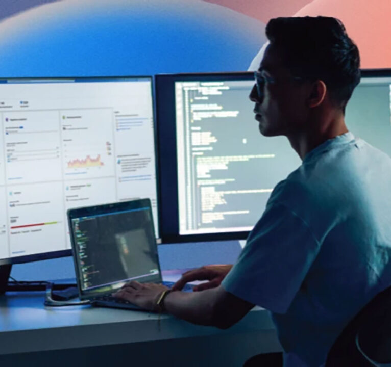 Man working on a laptop computer looking ahead at a large monitor screen.